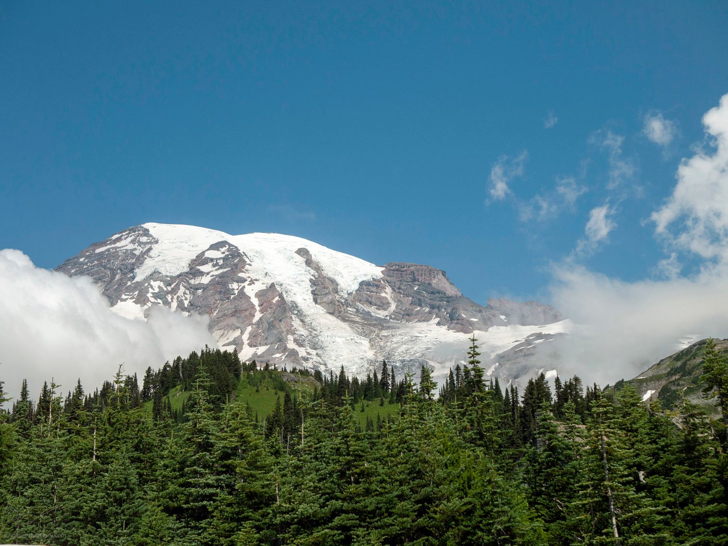 Landscape photo of the peak of Mt. Rainier, framed by clouds to the left and right, and trees in the foreground.
