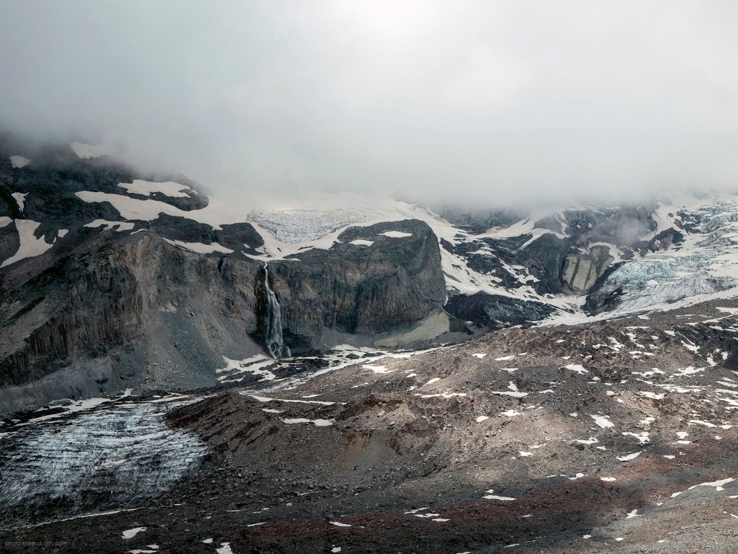 Landscape of a glacier. The top 1/3rd of the photo is clouds, and the rest is barren rocks and cliffs, spotted with snow