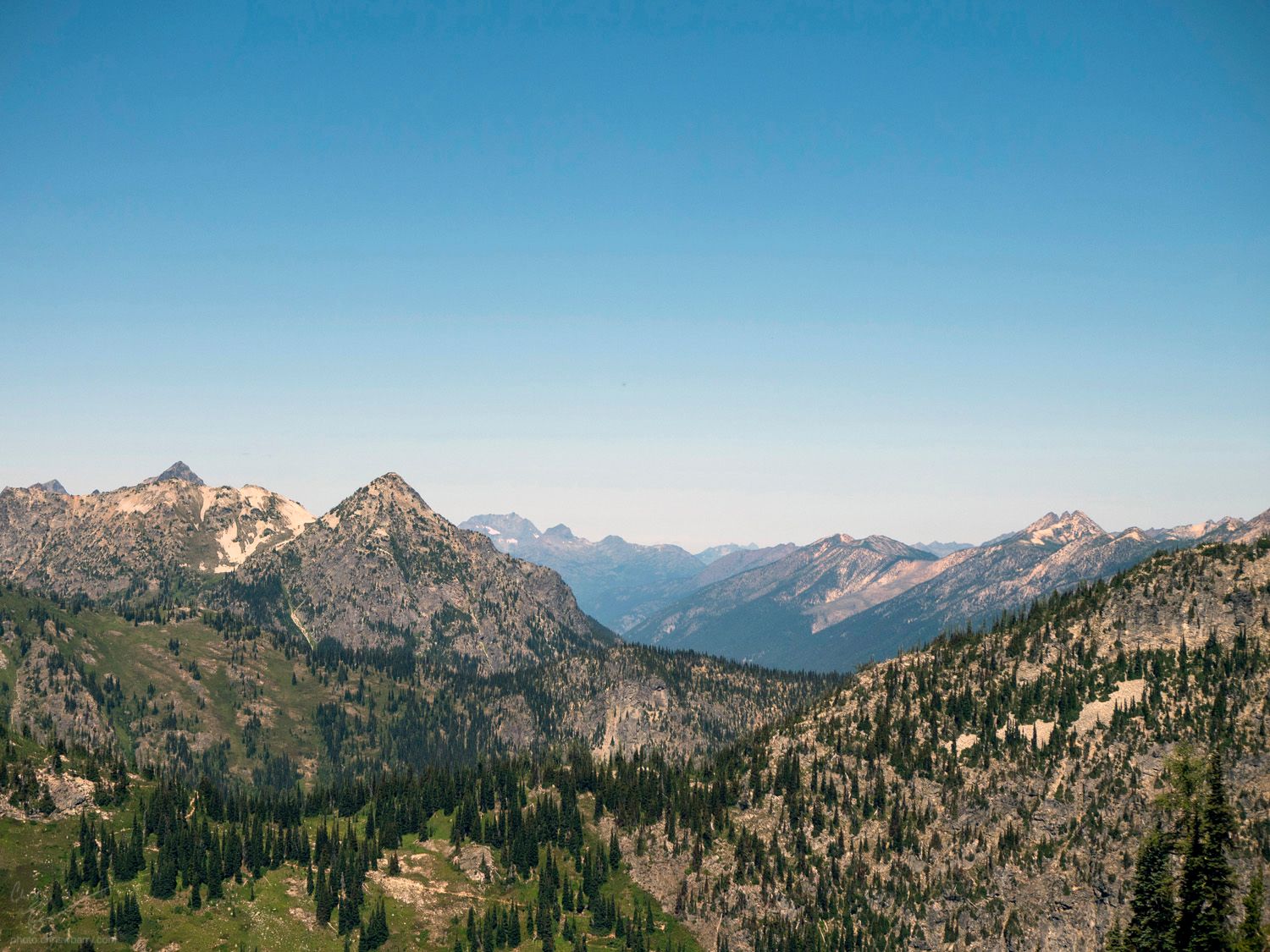 Landscape of Mountains dotted with pine trees, with a clear blue to pale white sky