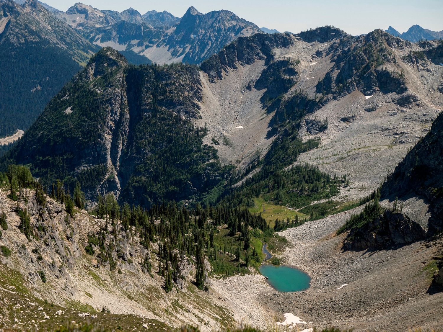 A large landscape of mountains, foregrounded with a small, blueish green lake, and dotted with patches of snow